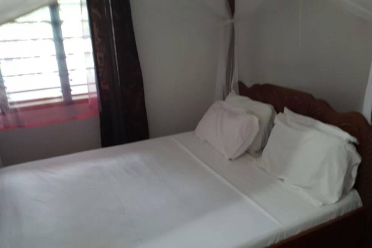 Una cama con sábanas blancas y cabecera de madera, una mesita de noche con lámpara, y una ventana con persianas.