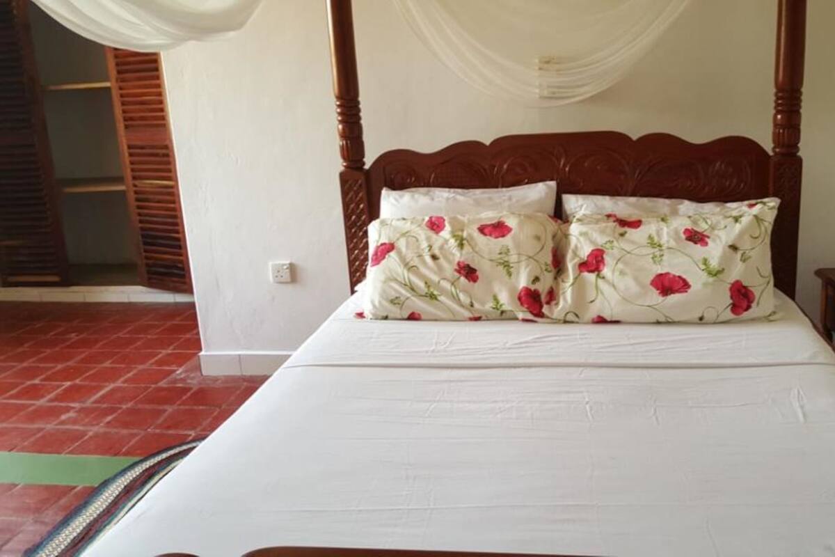 Una cama con ropa de cama blanca y almohadas florales, un cabecero de madera, una mesita de noche y un suelo de baldosas rojas.