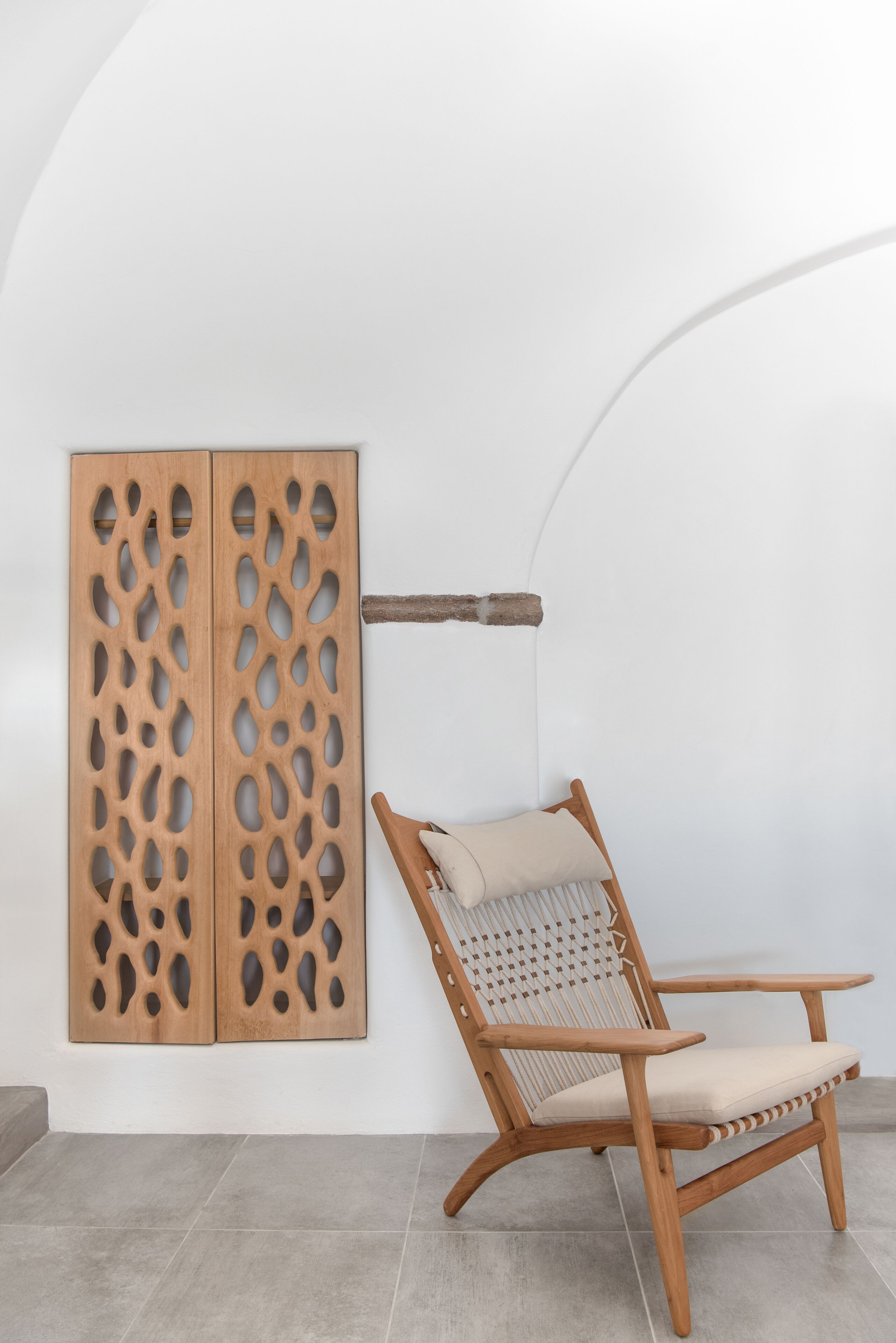 A wooden chair with a cushion, a wooden panel with cut-out patterns, and a simple white wall with a curved arch.