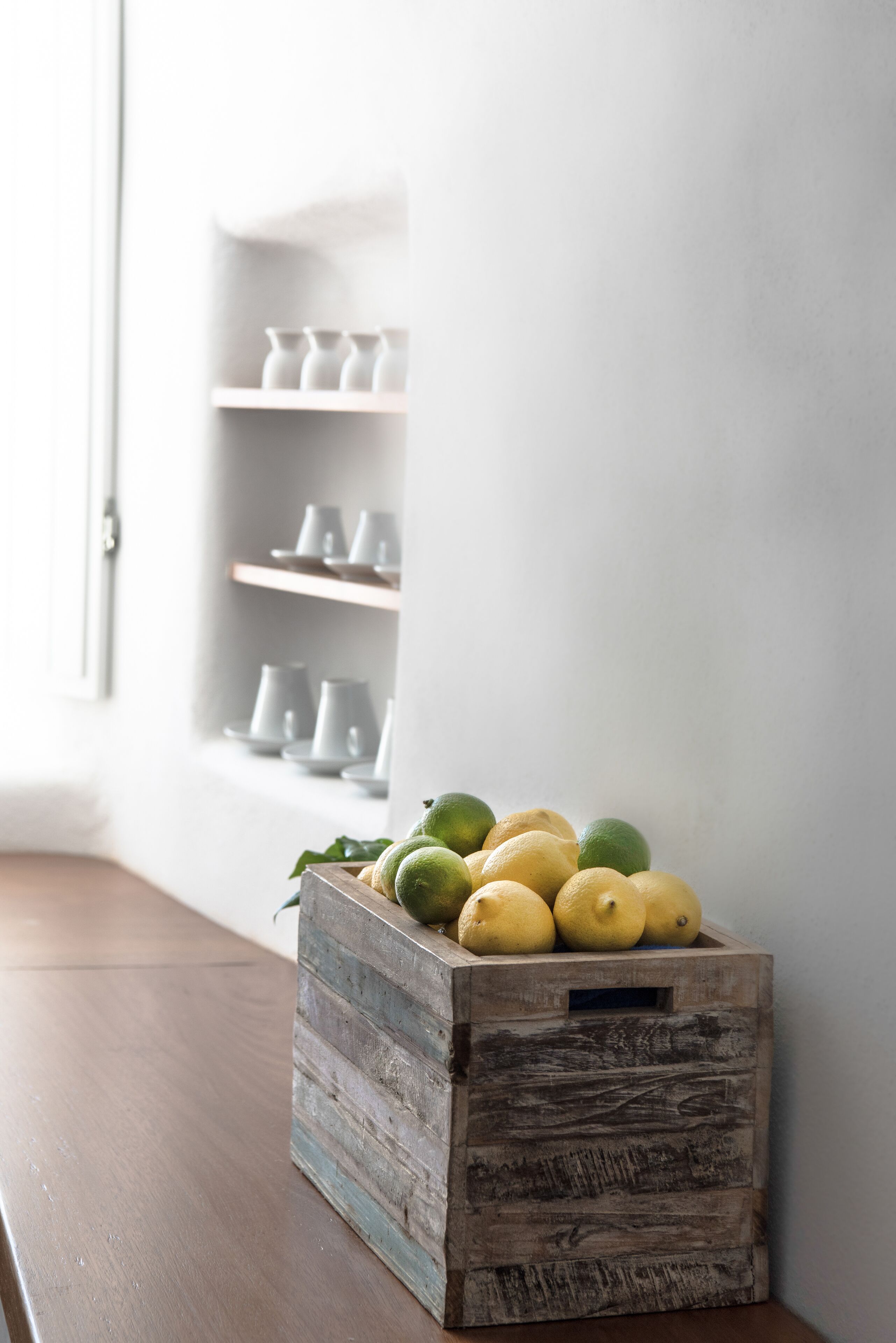 A wooden crate with lemons and limes on a wooden table, next to a white wall with shelves holding white cups.