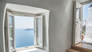 A room with a large window overlooking the sea, a wooden staircase, and a white door. - Native Eco Villas (Santorini)