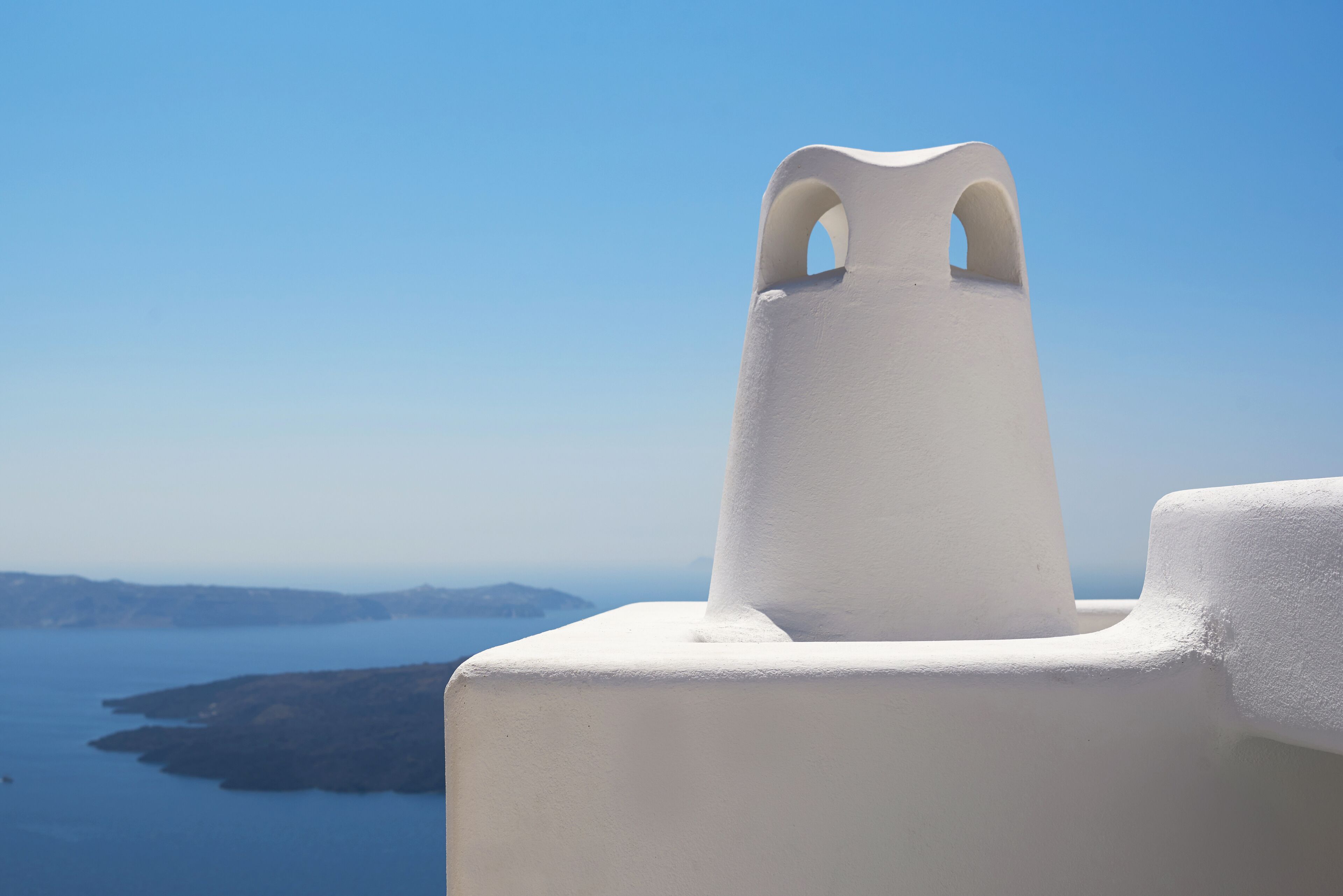 A white architectural structure with a prominent arched opening, overlooking a coastal landscape with a clear blue sky.