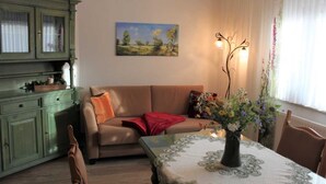 Un salon chaleureux avec un meuble vert, un canapé beige, une table recouverte d’une nappe à motifs floraux et un tableau représentant un paysage accroché au mur.