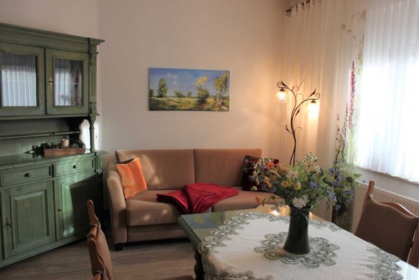 Un salon chaleureux avec un meuble vert, un canapé beige, une table recouverte d’une nappe à motifs floraux et un tableau représentant un paysage accroché au mur.
