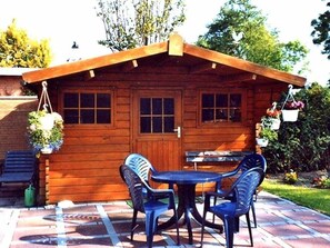 Eine Holzhütte mit Ziegelaußenwand, einer kleinen Terrasse mit Tisch und Stühlen sowie hängenden Blumenkörben.