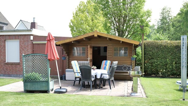 A wooden cabin with a patio, outdoor seating, and a hedge in the background. - Vacation house Langeoog, Baumann - Vacation house Baumann (Esens)