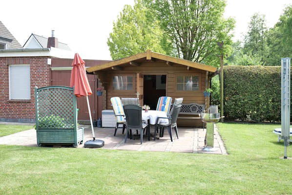 A wooden cabin with a patio, outdoor seating, and a hedge in the background.