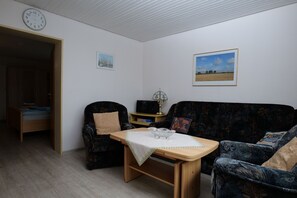 A living room with a wooden table, two armchairs, a sofa, a clock, and two framed pictures on the wall.