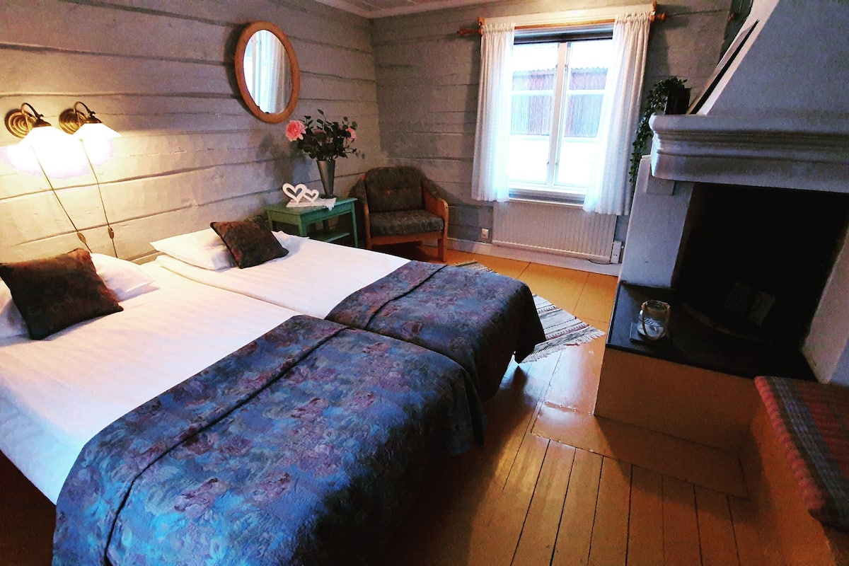 A room with two beds, a chair, a small table with a vase, a mirror, and a fireplace.
