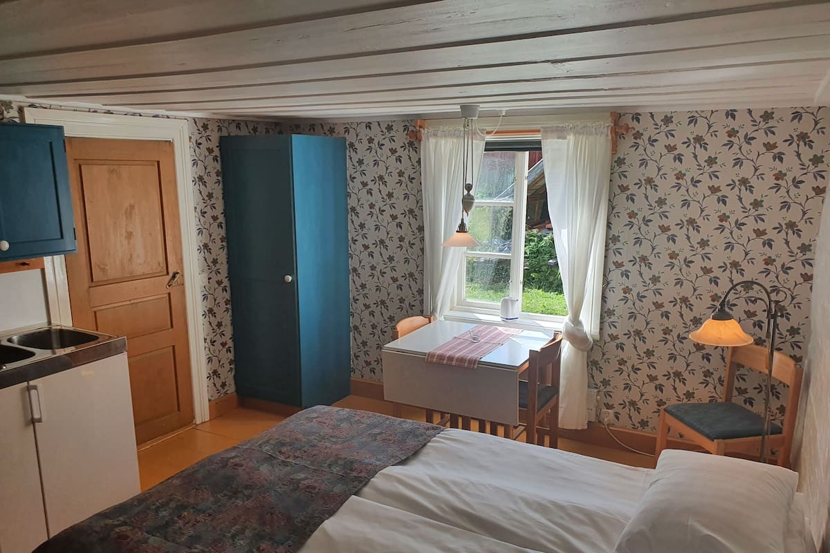 A compact bedroom with a bed, a small table with a chair, a window with curtains, and a kitchenette with a sink and cabinets.