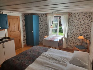 A compact bedroom with a bed, a small table with a chair, a window with curtains, and a kitchenette with a sink and cabinets. - Lövångergården (Lövånger)