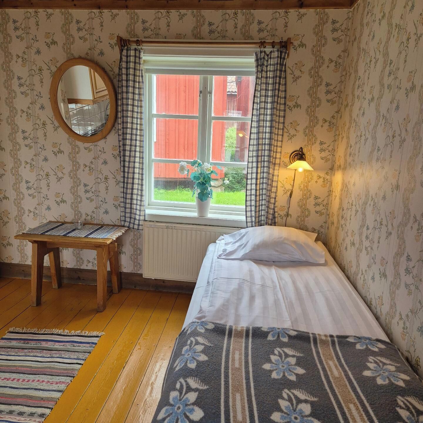 Une chambre à coucher comprenant un lit, une table en bois, un miroir, une fenêtre avec des rideaux, un radiateur et une lampe fixée au mur.