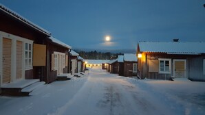 A snow-covered street lined with wooden buildings, a moon in the sky, and a clear view of the surrounding forest. - Lövångergården (Lövånger)