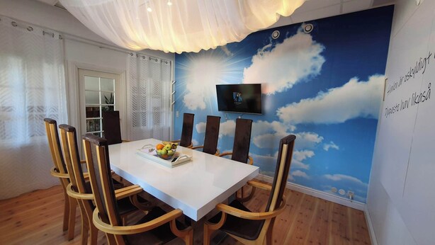 A dining area with a white table and wooden chairs, a TV mounted on the wall, and a mural of clouds and sun. - Lövångergården (Lövånger)