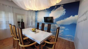A dining area with a white table and wooden chairs, a TV mounted on the wall, and a mural of clouds and sun. - Lövångergården (Lövånger)