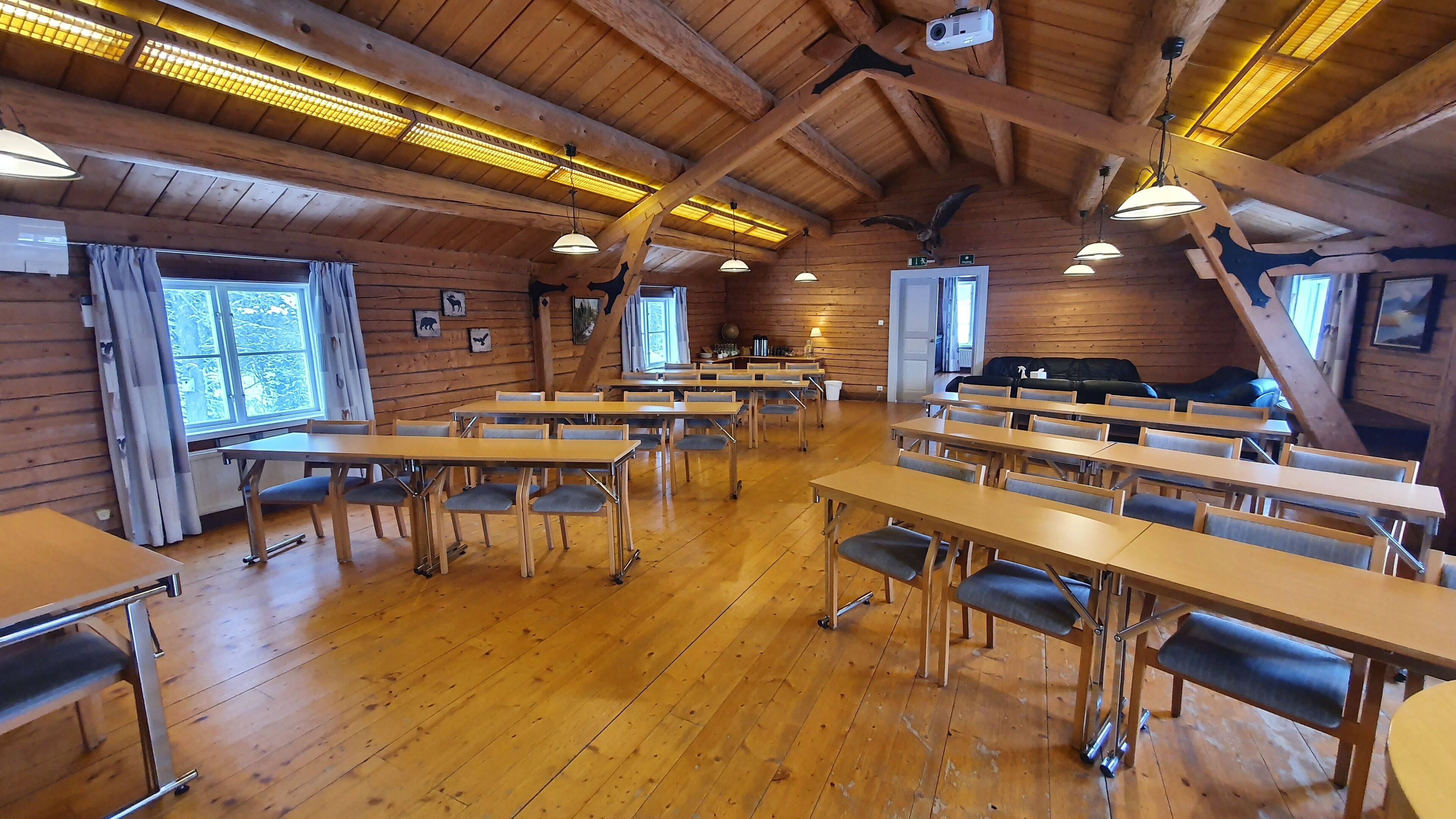 A spacious room with wooden walls and ceiling, equipped with tables and chairs, and a projector mounted on the ceiling.