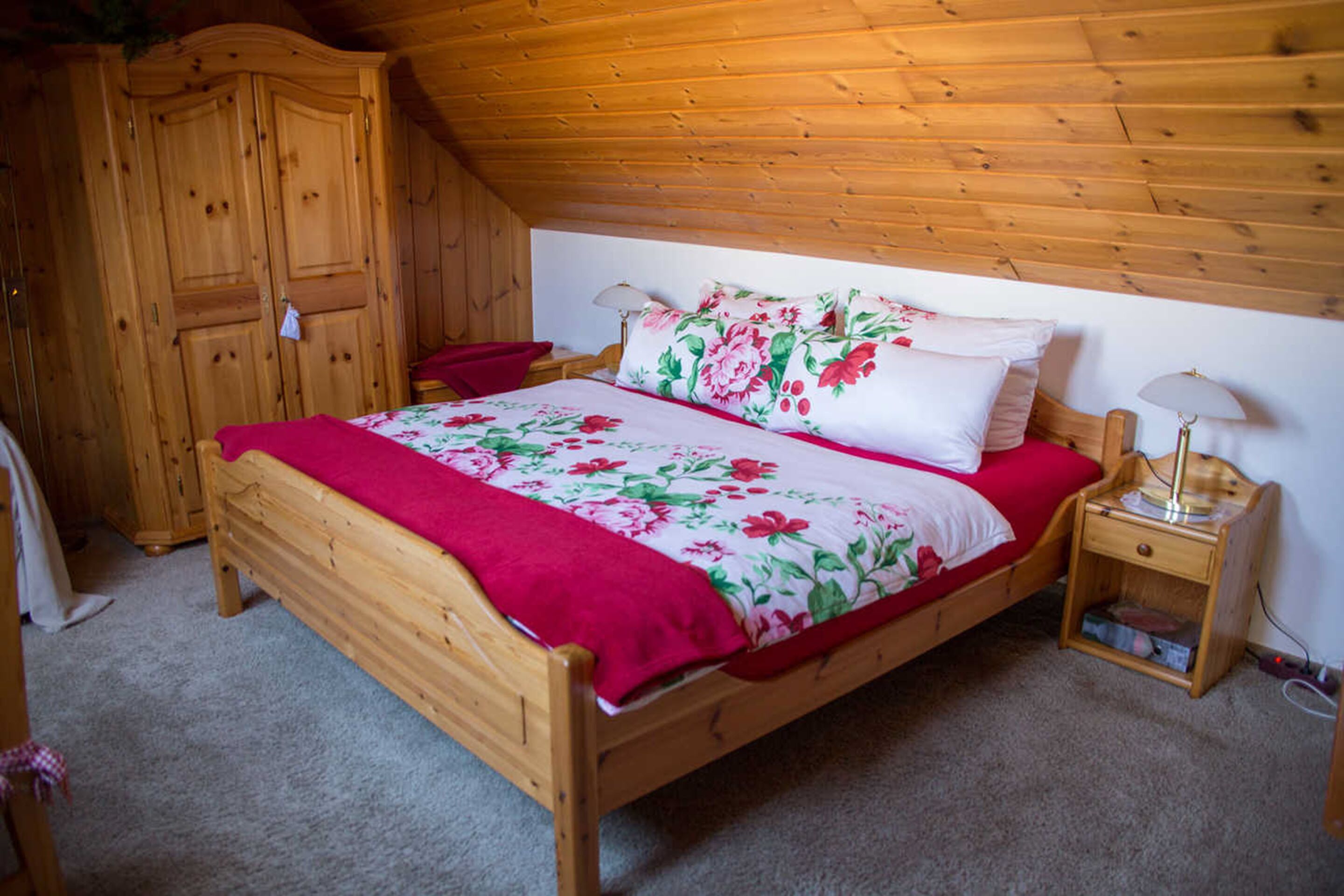 A wooden bed with a floral bedspread and red accents, a nightstand with a lamp, and a wooden wardrobe in a room with wooden ceiling beams.
