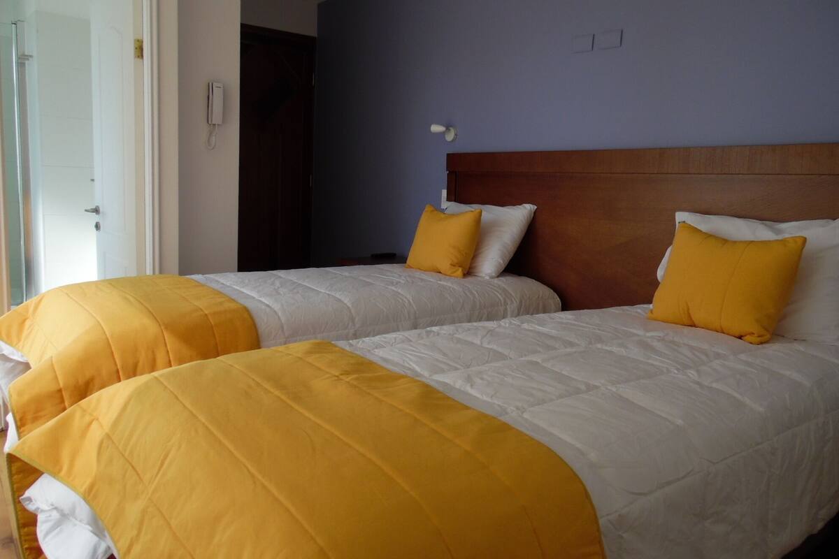 Hotellihuone, jossa on kaksi sänkyä. Jokaisessa sängyssä on keltaiset ja valkoiset vuodevaatteet, puinen pääty ja valkoinen ovi.