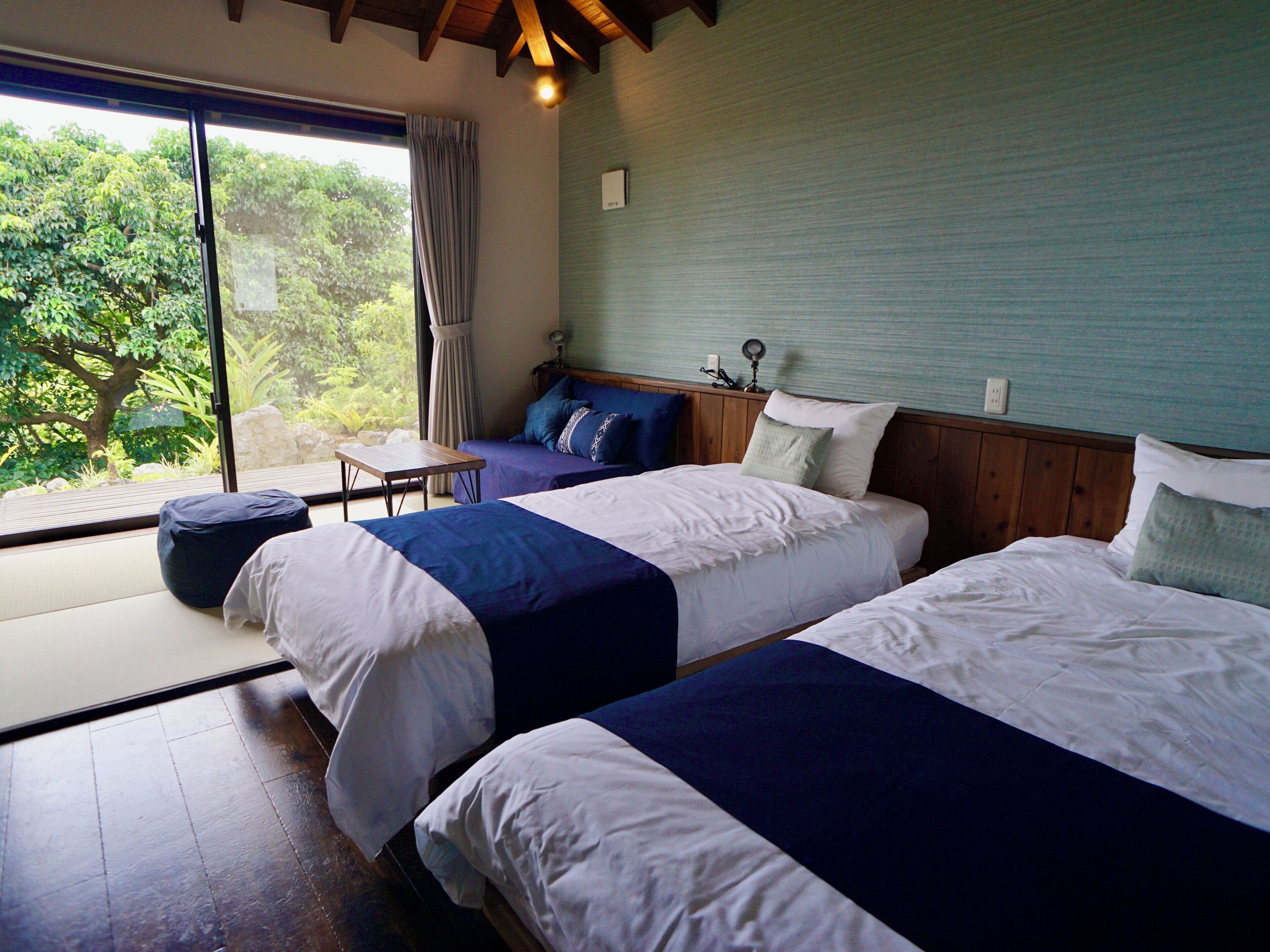 A room with two beds, a small table, and a view of greenery outside.