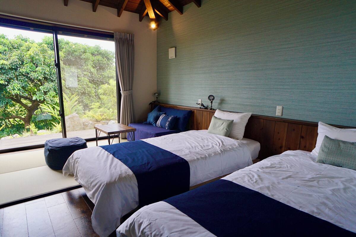 A room with two beds, a small table, and a view of greenery outside.