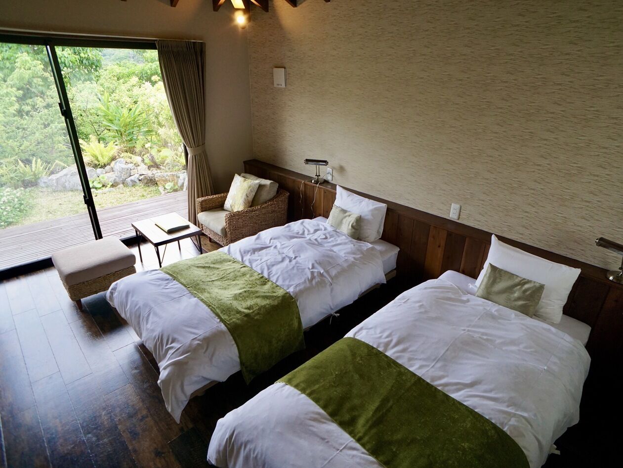 A hotel room with two beds, a small table, a chair, and a large window with a view of greenery.