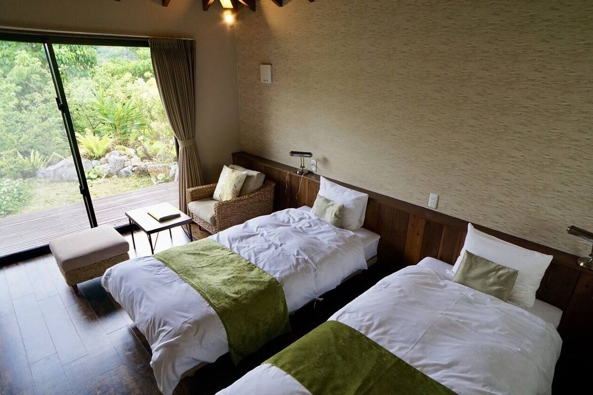 A hotel room with two beds, a small table, a chair, and a large window with a view of greenery.
