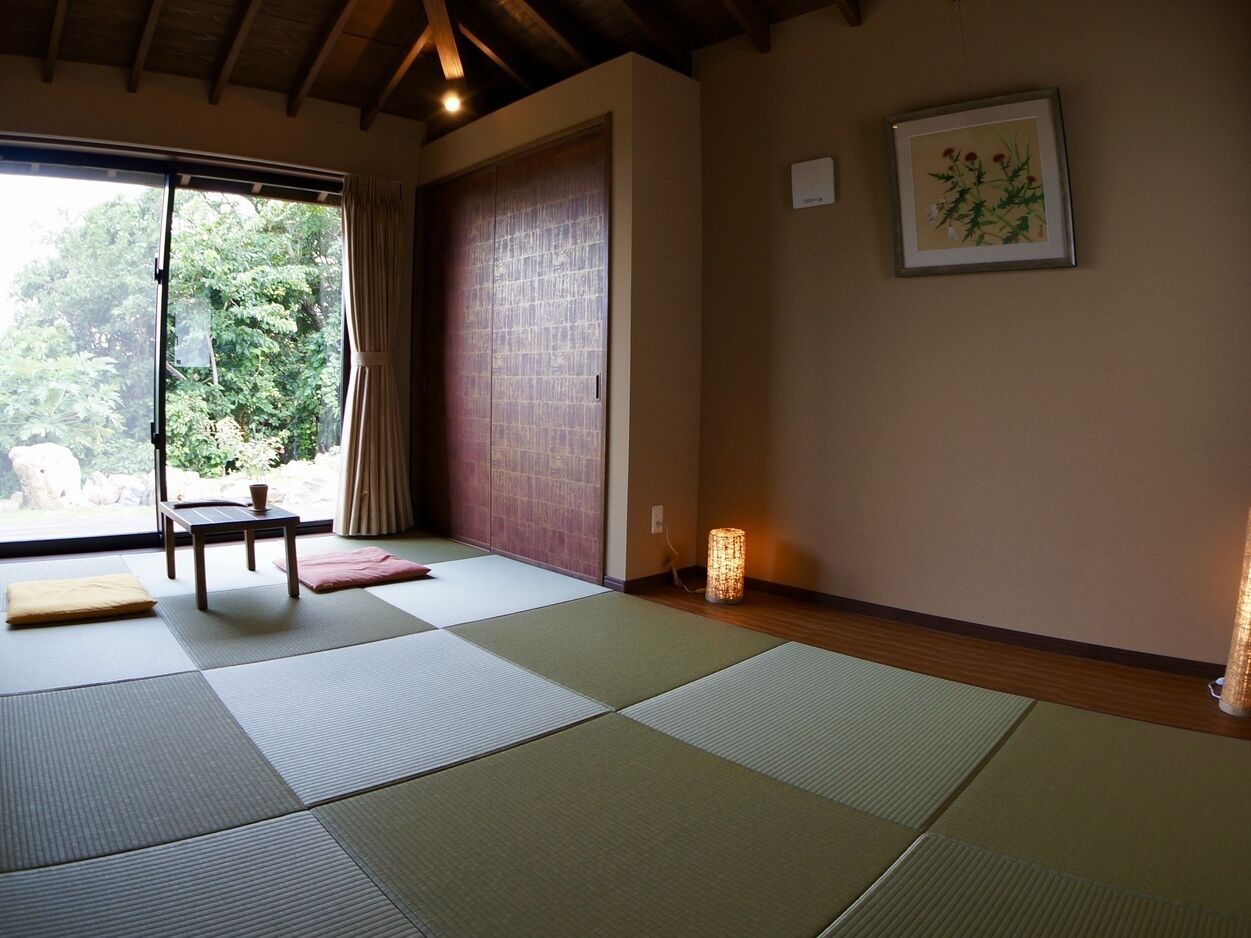 A room with tatami mats, a sliding door, a small table, a cushion, a lamp, and a framed picture on the wall.