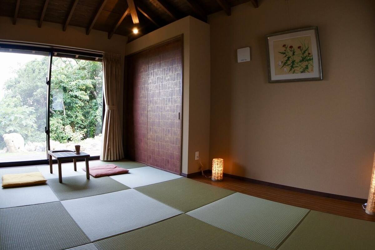 A room with tatami mats, a sliding door, a small table, a cushion, a lamp, and a framed picture on the wall.