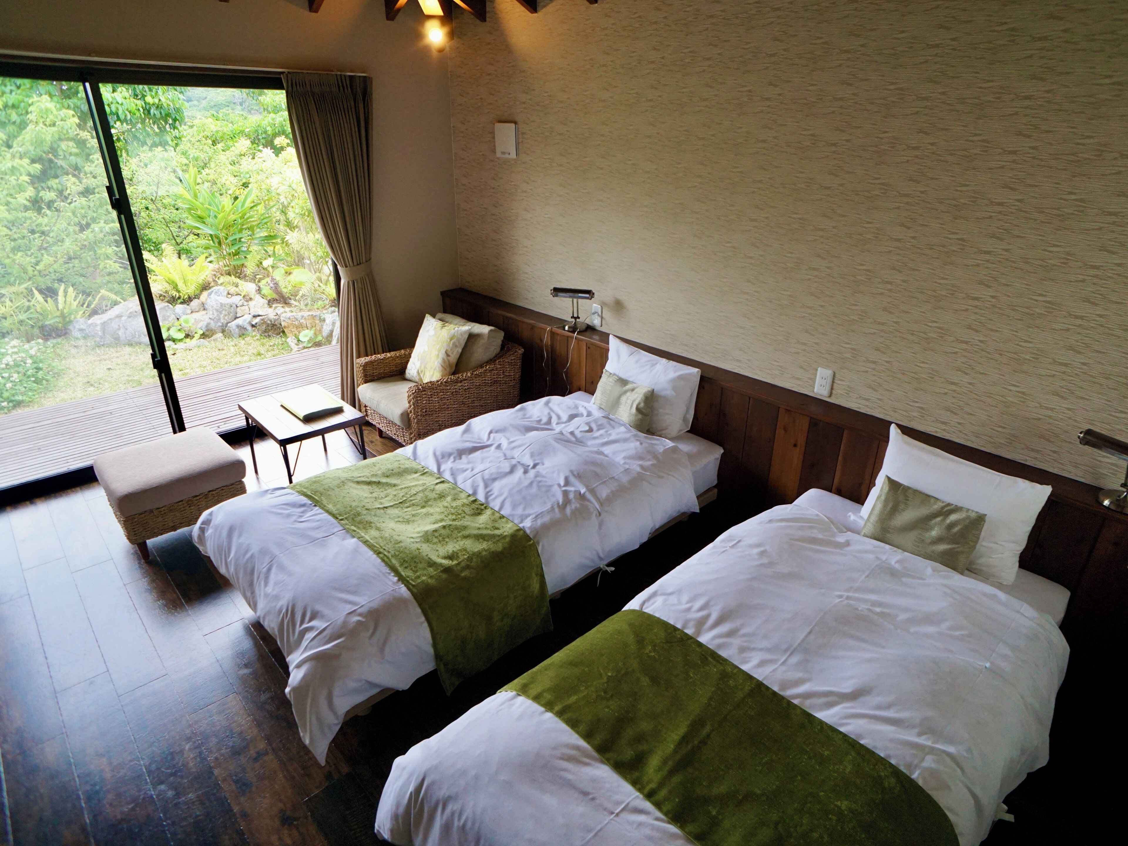 A hotel room with two beds, a small table, a chair, and a view of greenery outside.