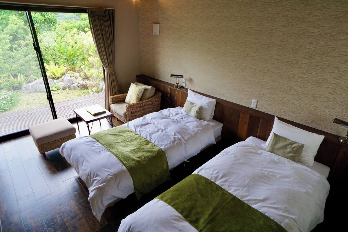A hotel room with two beds, a small table, a chair, and a view of greenery outside.