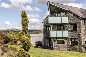 A two-story stone house with a balcony, surrounded by a well-maintained garden and a clear sky.