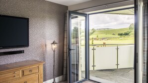 A room with a wooden dresser, a floor lamp, and a sliding glass door leading to a balcony with a view of a green landscape. - The Penthouse @ Carus Green (Kendal)