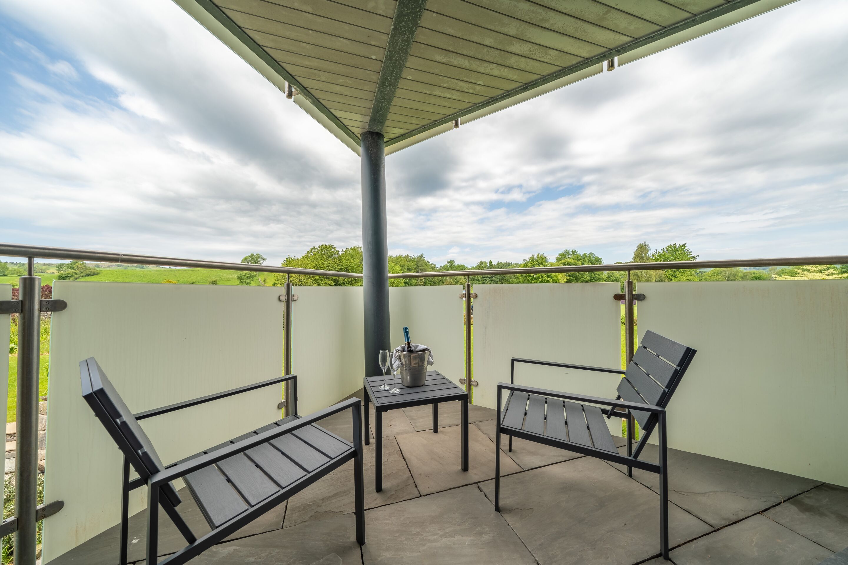Eine Terrassenanlage im Freien mit zwei Metallstühlen, einem Tisch, einem Metallpfosten und Blick auf ein grünes Feld unter einem bewölkten Himmel.