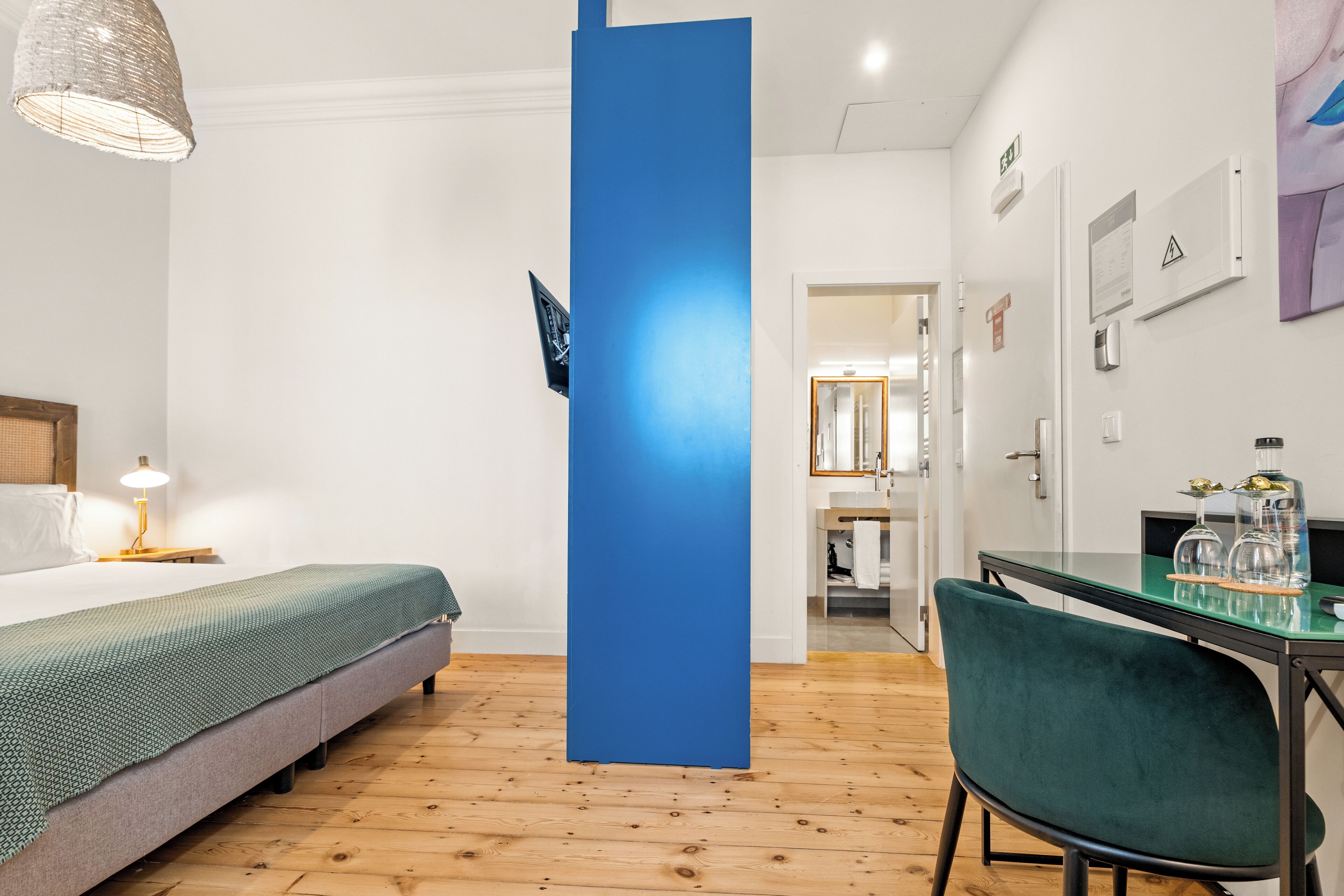 Quarto de hotel moderno com cama, escrivaninha com cadeira, parede azul decorativa e piso de madeira.