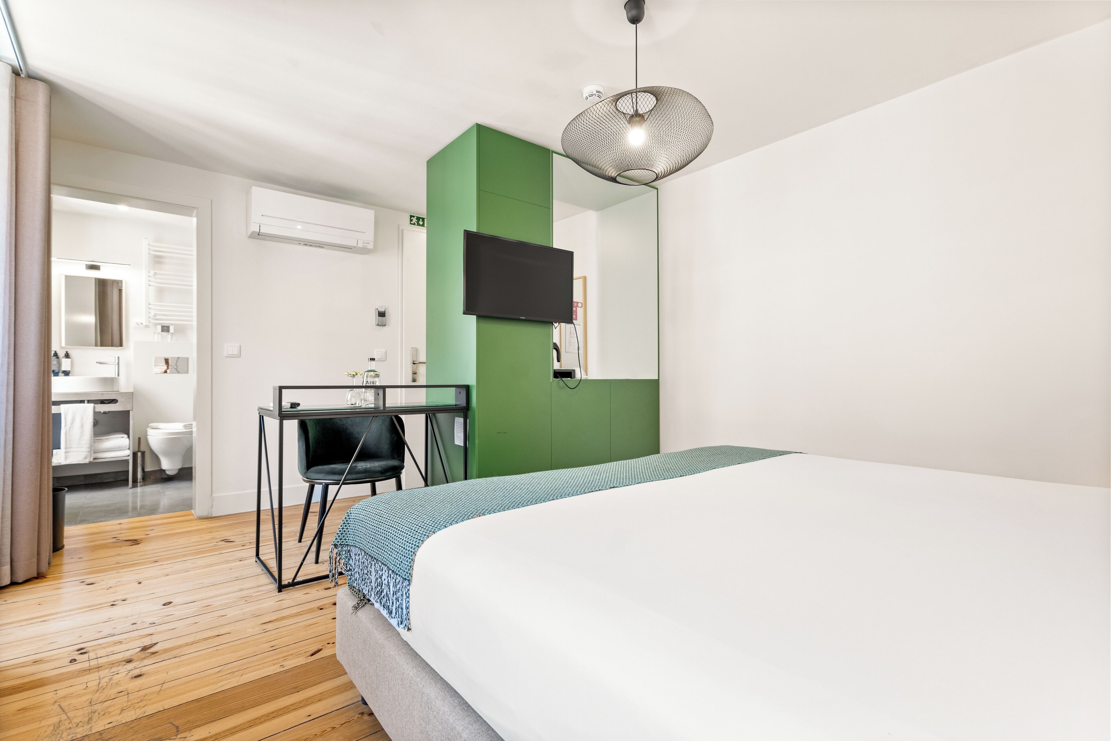 Une chambre d’hôtel moderne avec un plancher en bois, un lit recouvert d’une courtepointe blanche et bleue, un bureau avec une chaise, un téléviseur fixé à un mur vert et un luminaire suspendu.