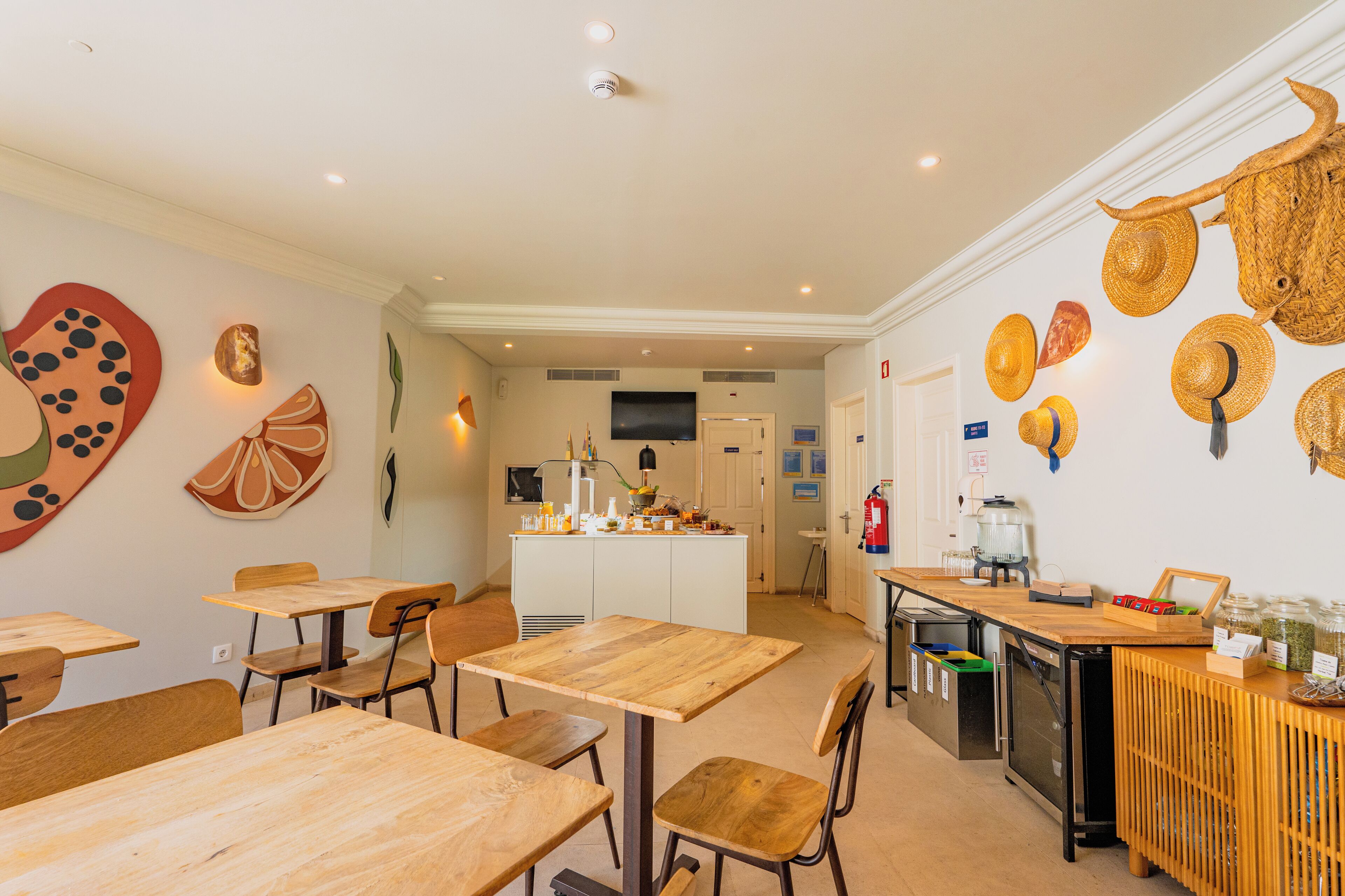 Un comedor con mesas y sillas de madera, una encimera de cocina con diversos objetos, y adornos de pared que incluyen sombreros y rodajas de fruta.