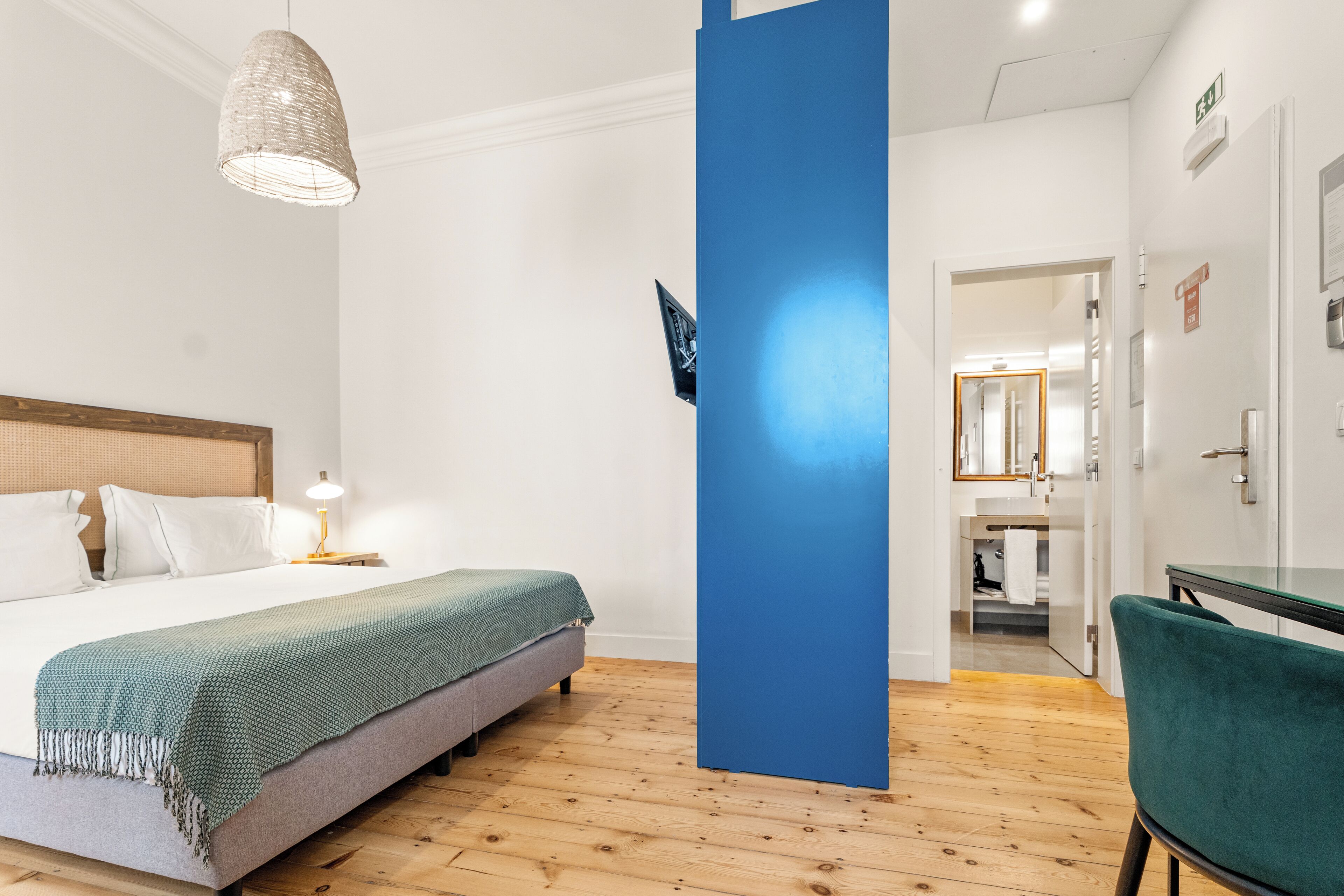 A hotel room with a bed, a desk, a chair, a blue pillar, and a hanging light fixture.
