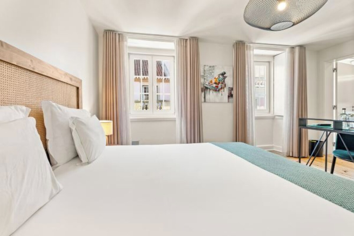 A hotel room with a large bed, a wooden headboard, a window with curtains, a small table with a chair, and a painting on the wall.