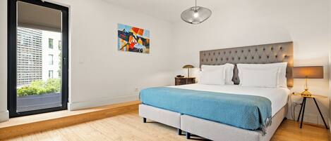 A modern bedroom with a large bed, a window with a view of buildings, a painting on the wall, and a ceiling light.
