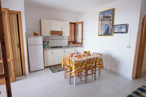 Cocina compacta con una mesa de comedor lista para cuatro personas, un refrigerador y un ventanal con persianas.