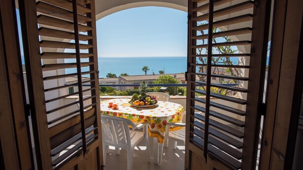An open wooden door with shutters leading to a balcony with a table set for two, overlooking a coastal view. - Holiday House With Panoramic View, Balcony, Wifi, and Near the Sea (Valderice)