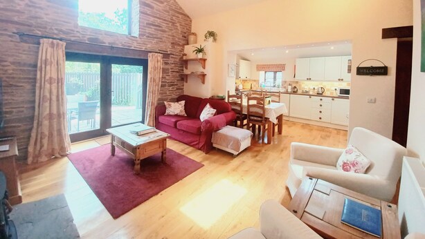A spacious living room with a stone wall, a red sofa, a wooden coffee table, and a white armchair. A kitchen is visible in the background with white cabinets and a welcome sign. - A peaceful escape in rural Herefordshire - perfect for families and friends. (Nr. Hereford)