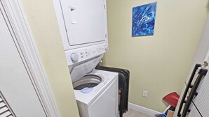 A laundry room with a front-loading washer and dryer, a painting on the wall, and a red trash bin. - AD 1 2104, a spacious 2-bedroom condo in Destin, Fabulous views (Destin)