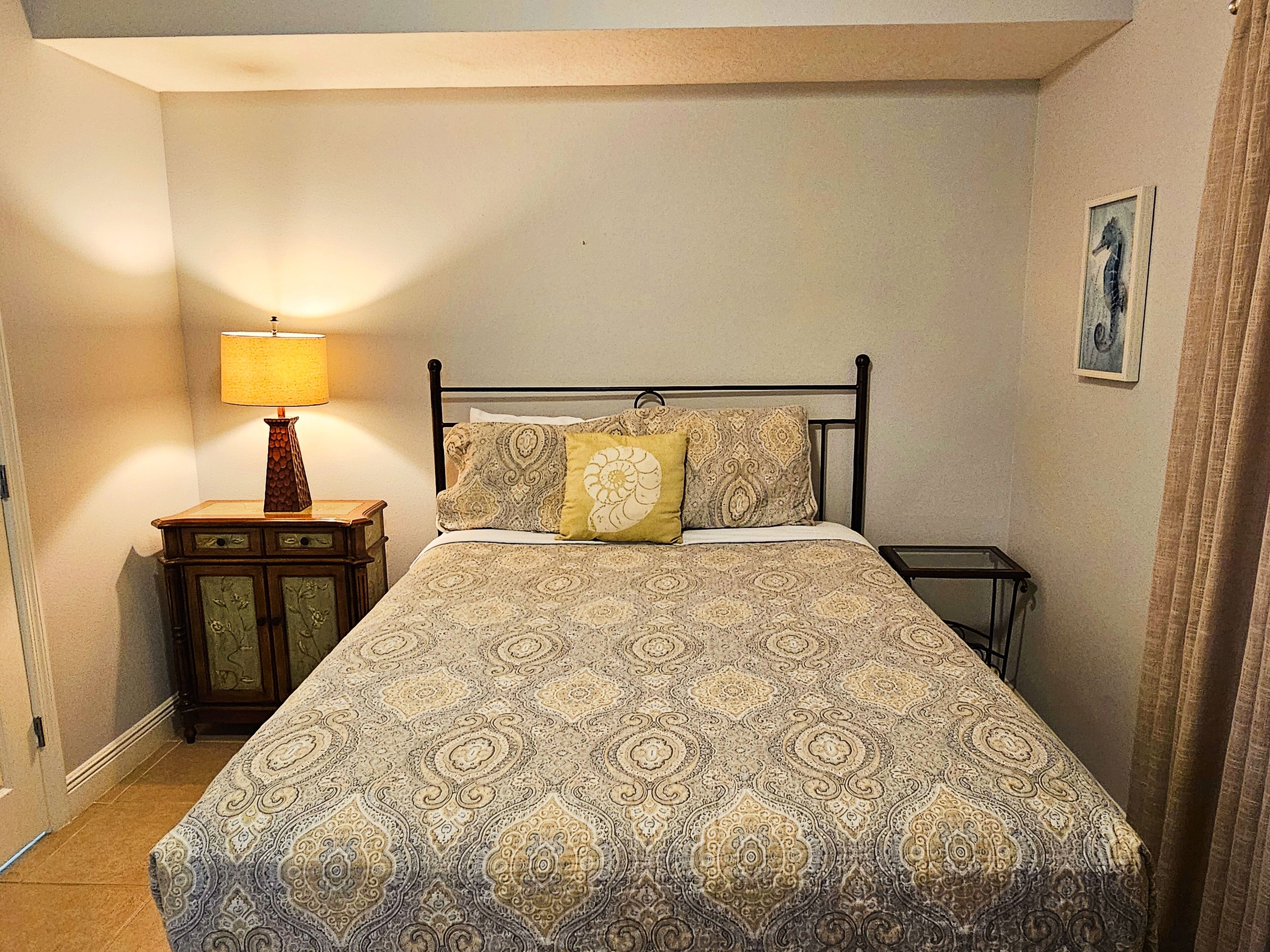 A neatly made bed with a patterned bedspread and two pillows, a bedside table with a lamp, and a framed picture on the wall.