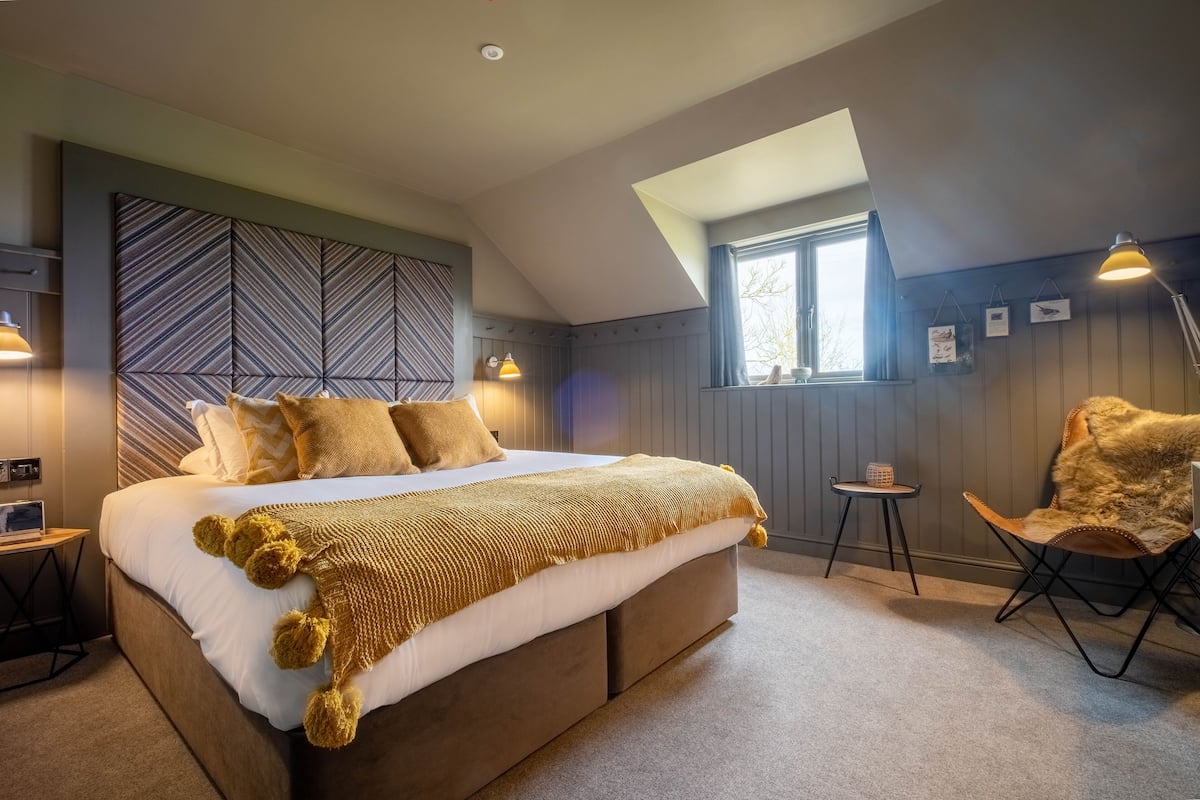 Une chambre avec un grand lit, une tête de lit ornée d’un motif géométrique, une petite table avec une lampe et une chaise recouverte d’un plaid en fausse fourrure.