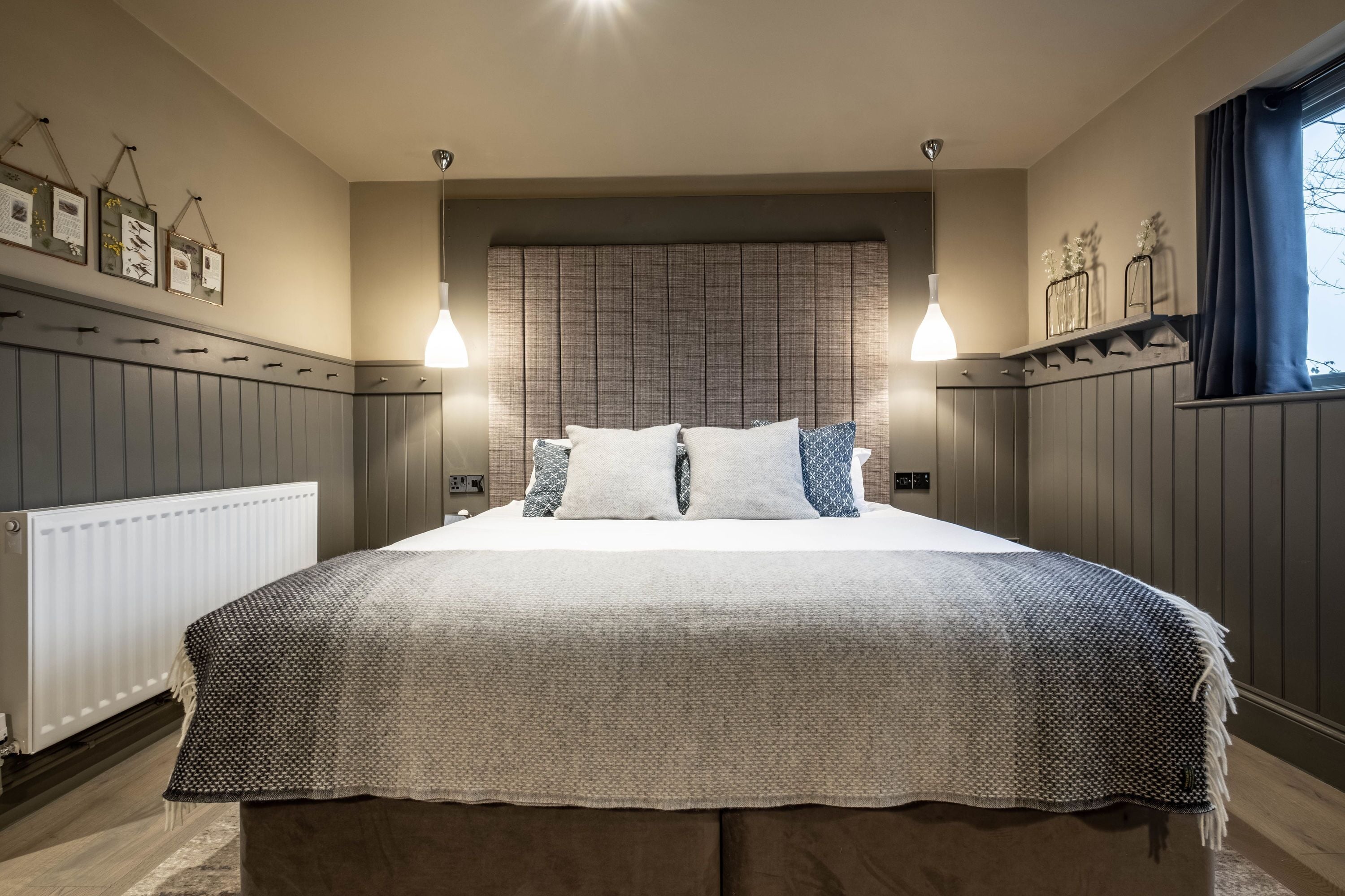A neatly arranged bedroom with a large bed, two bedside lamps, a radiator, and wall-mounted shelves with decorative items.