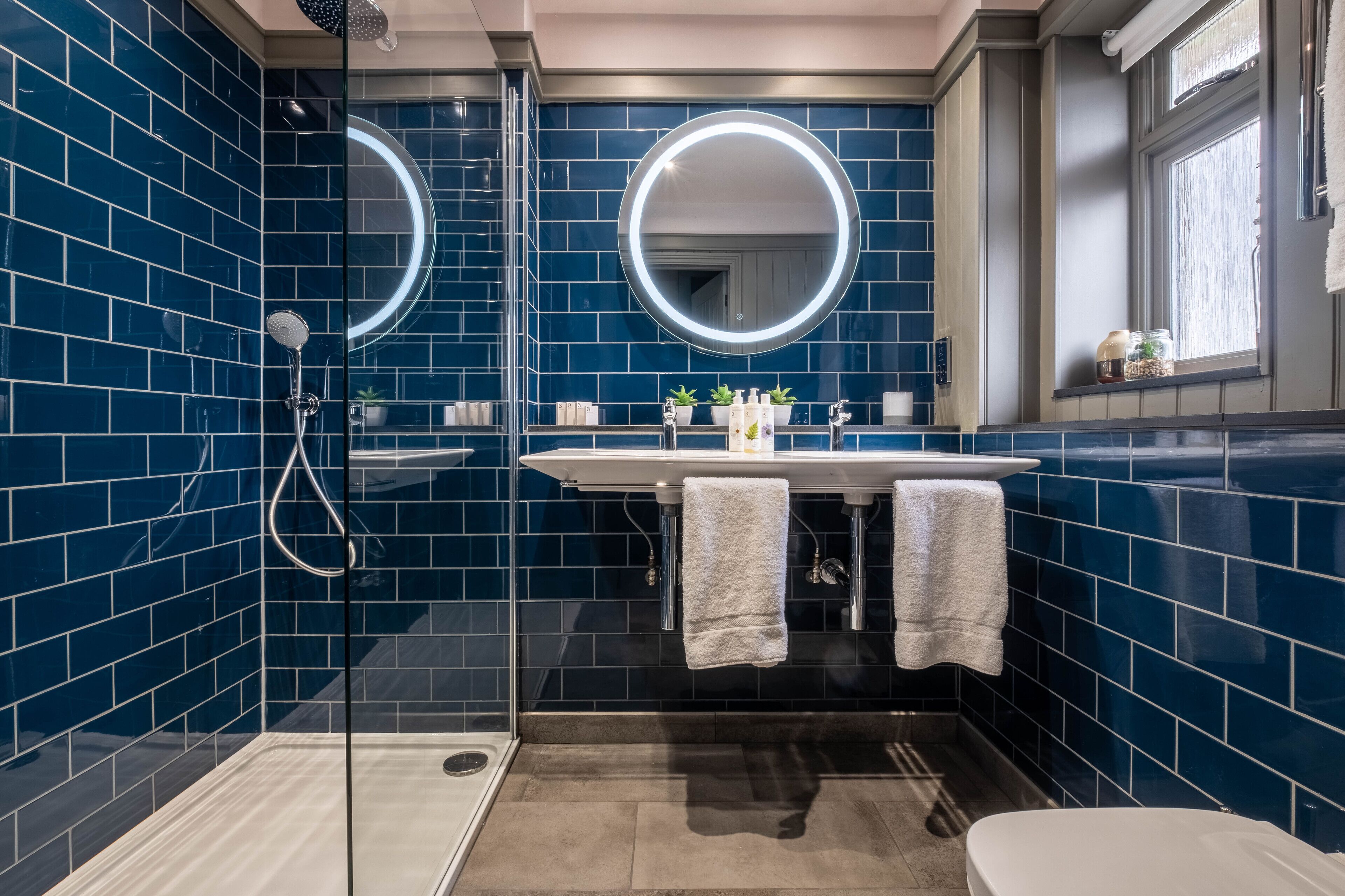 A modern bathroom with blue tiles, a glass shower enclosure, a white sink with two towels, and a round mirror with a light.