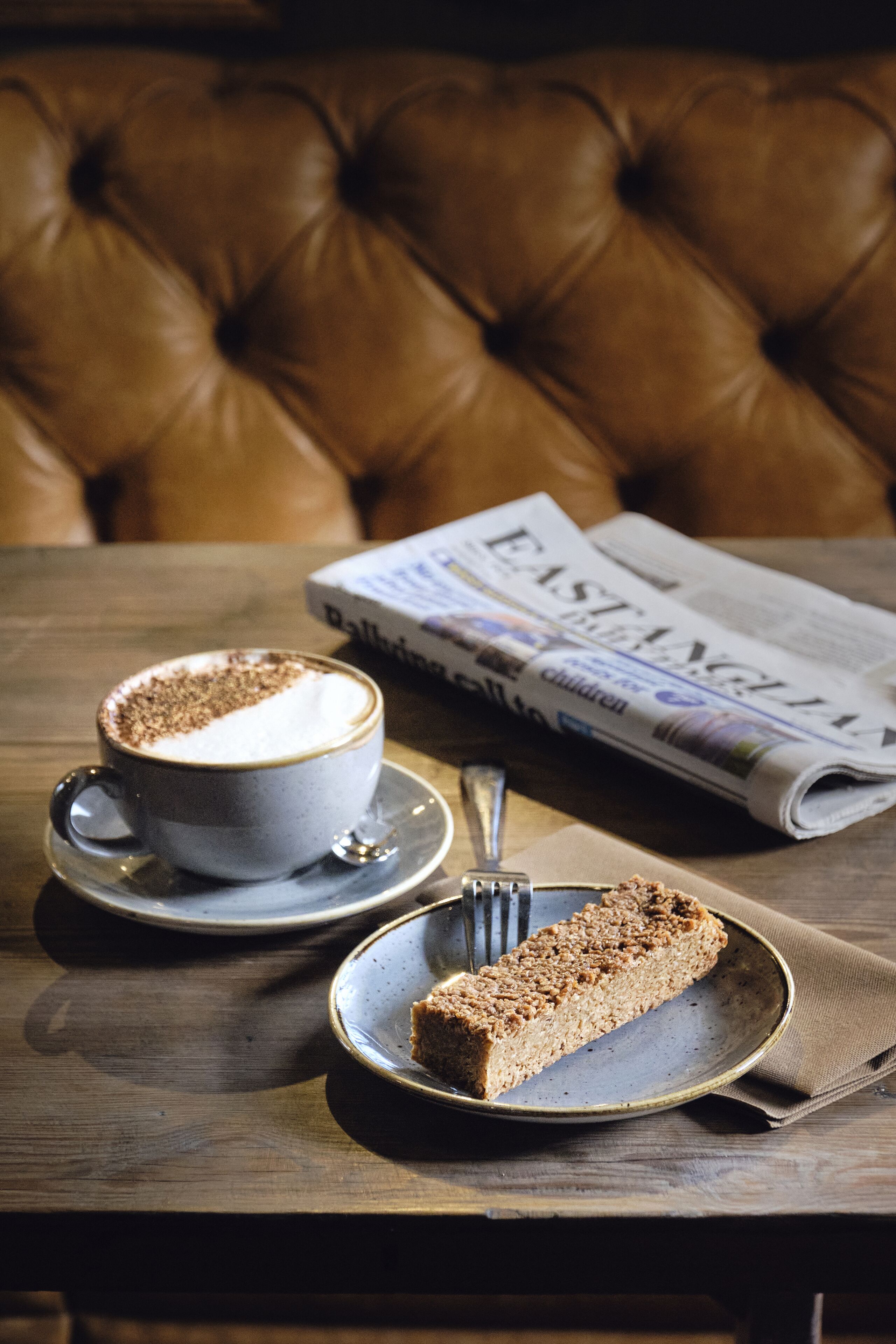 Uma mesa com uma xícara de café, uma fatia de bolo e um jornal.