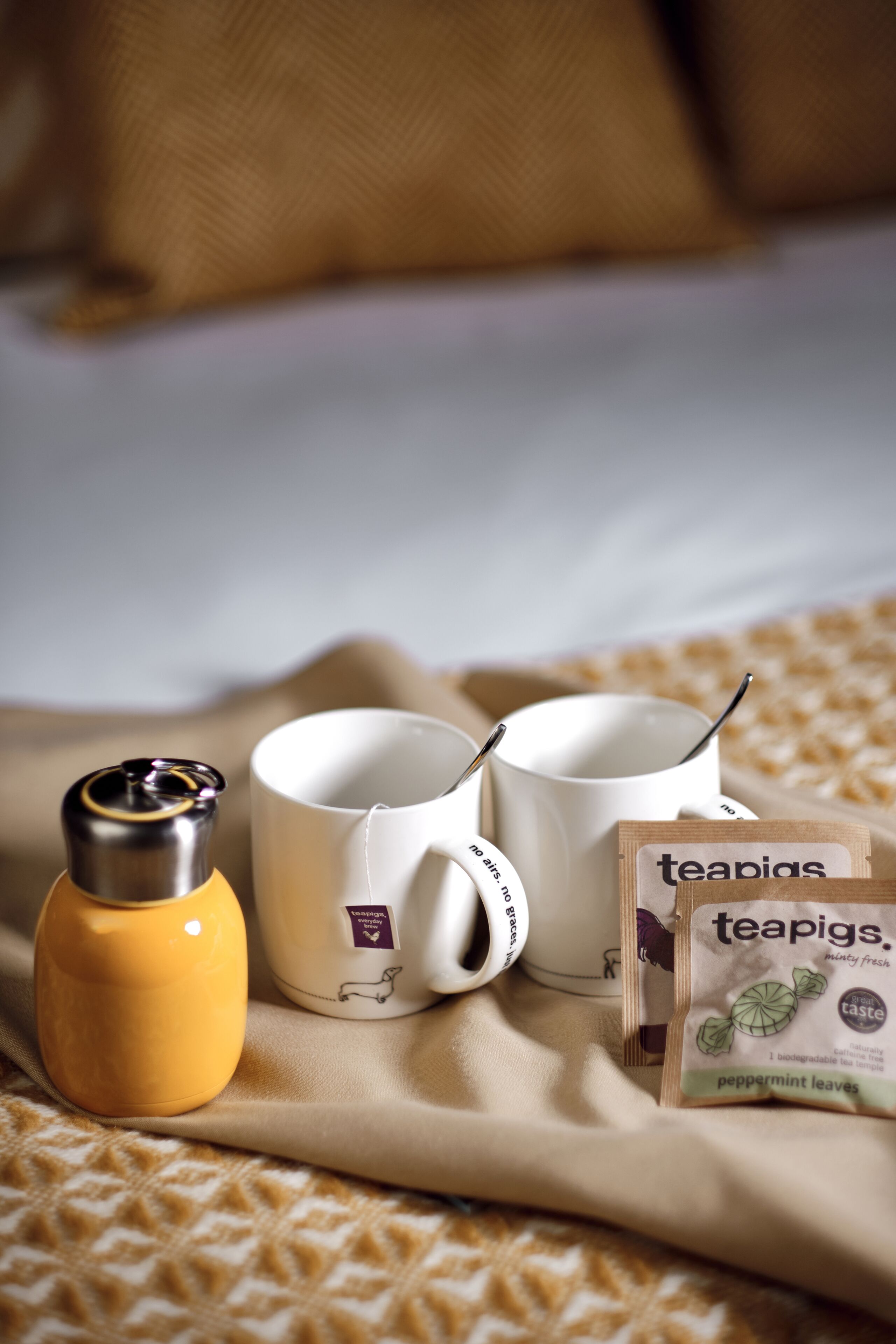 A bed with a beige blanket, two white mugs, a yellow thermos, and a package of teapigs peppermint leaves.