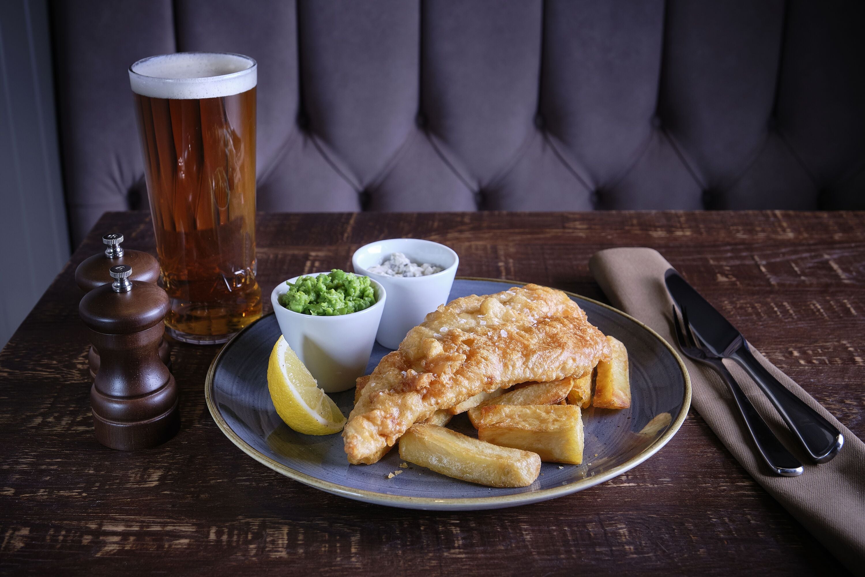 Un plato de pescado con papas fritas, una cerveza y salsas sobre una mesa de madera.